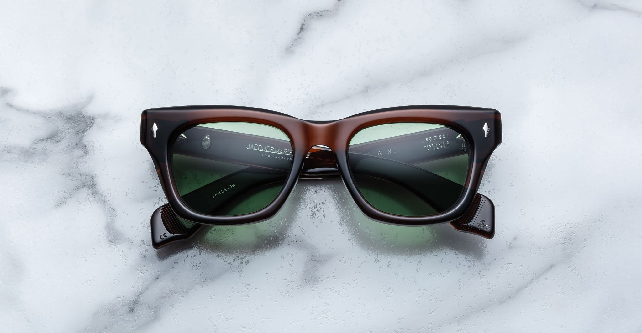 The Jacques Marie Mage Dealan sunglasses, featuring rectangular dark frames and green tinted lenses, are shown on a white marble surface. White printed text and branding appear inside the arms.