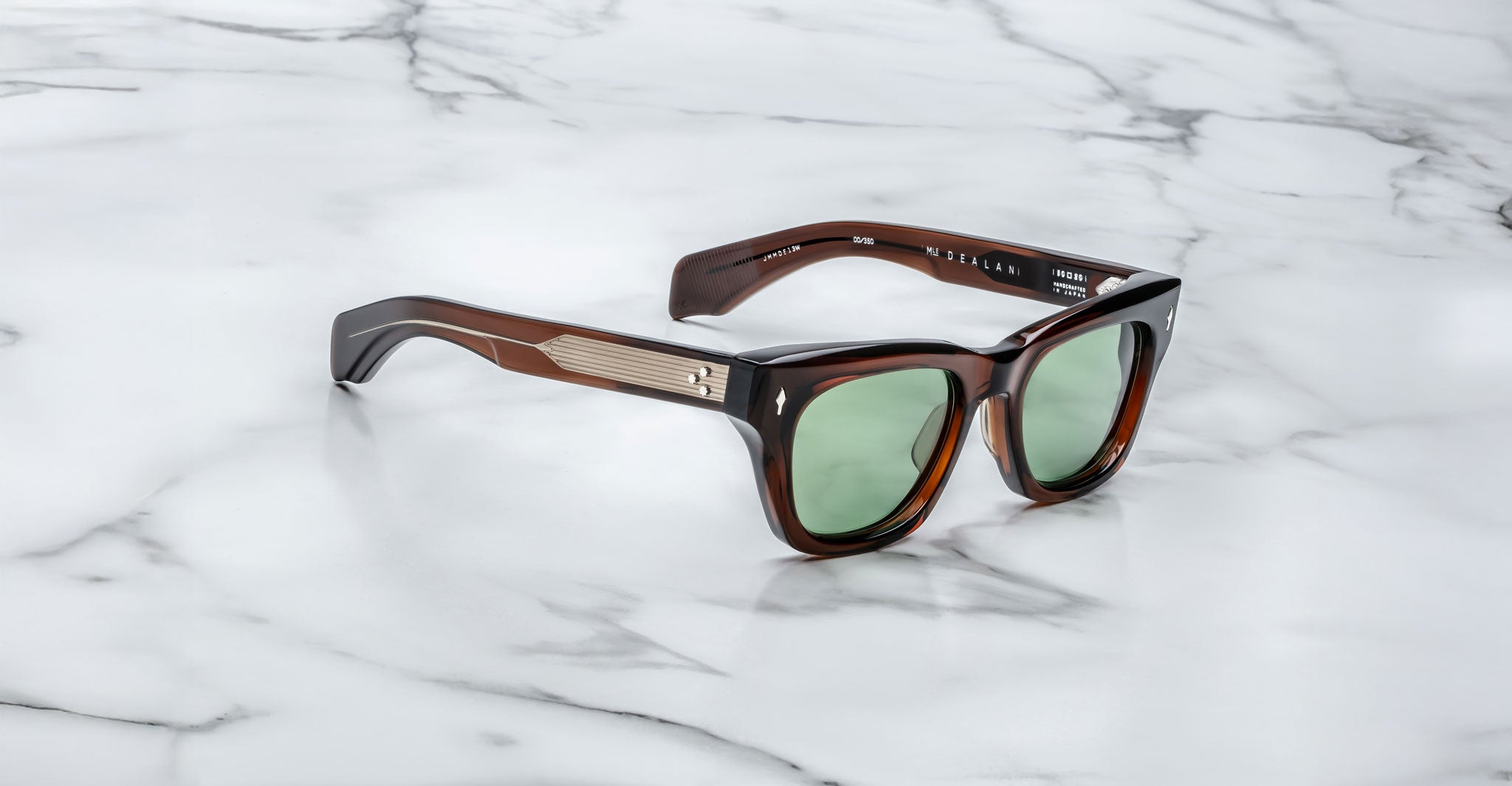 The Jacques Marie Mage Dealan sunglasses feature dark brown frames, green-tinted lenses, and silver accents on thick arms, displayed on a white marble surface.
