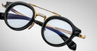 A close-up of Jacques Marie Mage Delage eyeglasses featuring round lenses, black frames with gold accents, and a distinctive nose bridge design set against a light gray background.