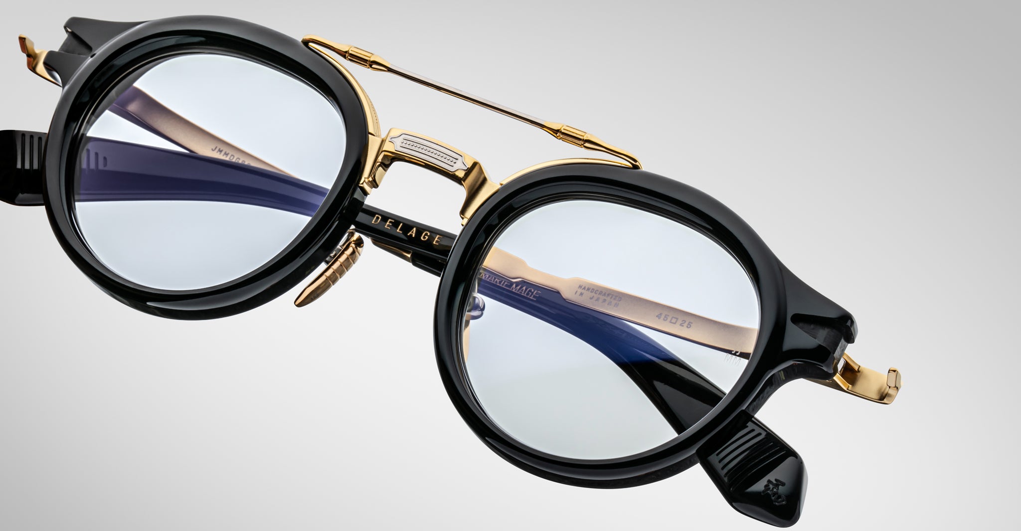 A close-up of Jacques Marie Mage Delage eyeglasses featuring round lenses, black frames with gold accents, and a distinctive nose bridge design set against a light gray background.