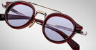 Close-up of Jacques Marie Mage Delage eyeglasses featuring round, dark red frames, purple-tinted lenses, gold accents on the nose bridge and nose pads, with brand markings on the inner arms, set against a plain background.