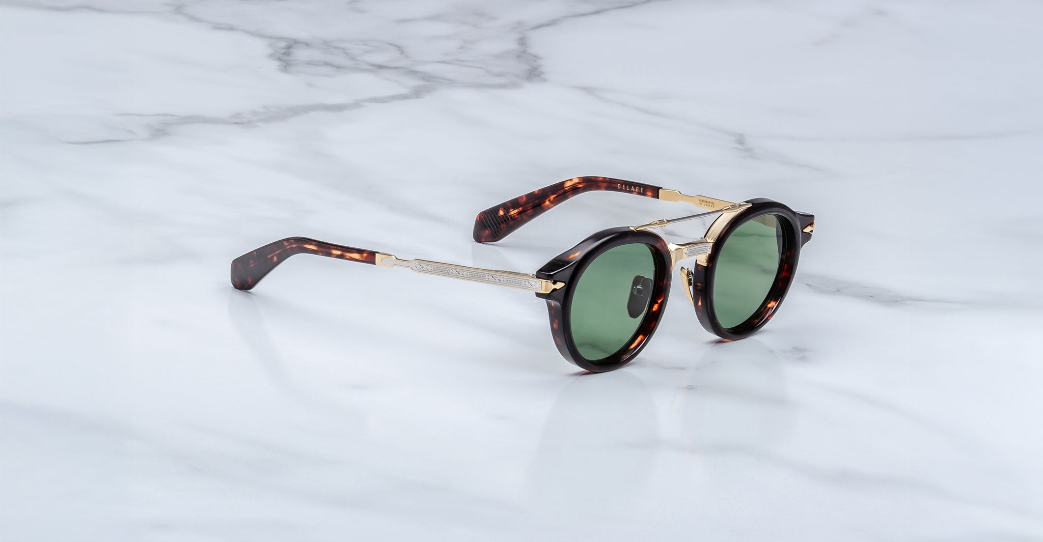 The Jacques Marie Mage Delage sunglasses feature round green lenses, a tortoiseshell frame, and gold metal temples, shown on a white marble surface.