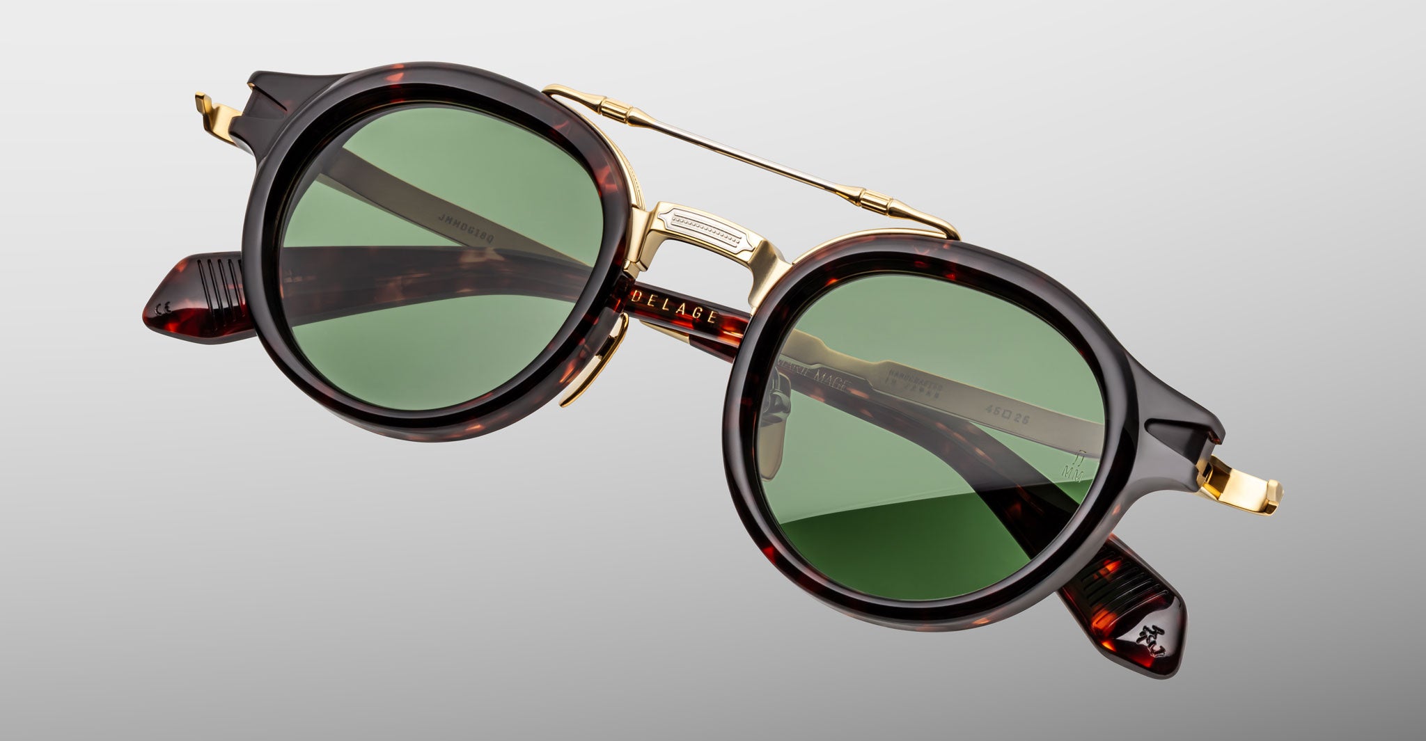 The Jacques Marie Mage Delage sunglasses feature round green lenses, a dark tortoiseshell frame, and gold metal accents with a double bridge and hinges, shown against a light gray background.