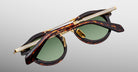A pair of Jacques Marie Mage Delage tortoiseshell sunglasses with green lenses is folded, revealing detailed gold accents and engraved patterns on the arms and bridge against a plain gray background.