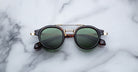 The Jacques Marie Mage Delage sunglasses, featuring round dark green lenses, a tortoiseshell frame with gold accents on the hinges and nose bridge, and a gold bridge, are unfolded on a white marble surface.
