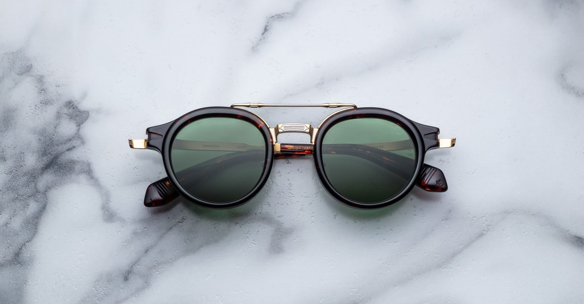 The Jacques Marie Mage Delage sunglasses, featuring round dark green lenses, a tortoiseshell frame with gold accents on the hinges and nose bridge, and a gold bridge, are unfolded on a white marble surface.