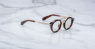 Jacques Marie Mage Delage round tortoiseshell eyeglasses with gold metal temples and bridge, displayed open on a white marble surface.