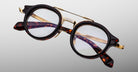 The Jacques Marie Mage Delage is a pair of round eyeglasses featuring tortoiseshell acetate frames, gold metal bridge and temples, and clear lenses, shown against a light gray background.