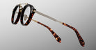 The Jacques Marie Mage Delage features round eyeglasses with thick, dark tortoiseshell frames and gold metal temples, shown against a gray gradient background.
