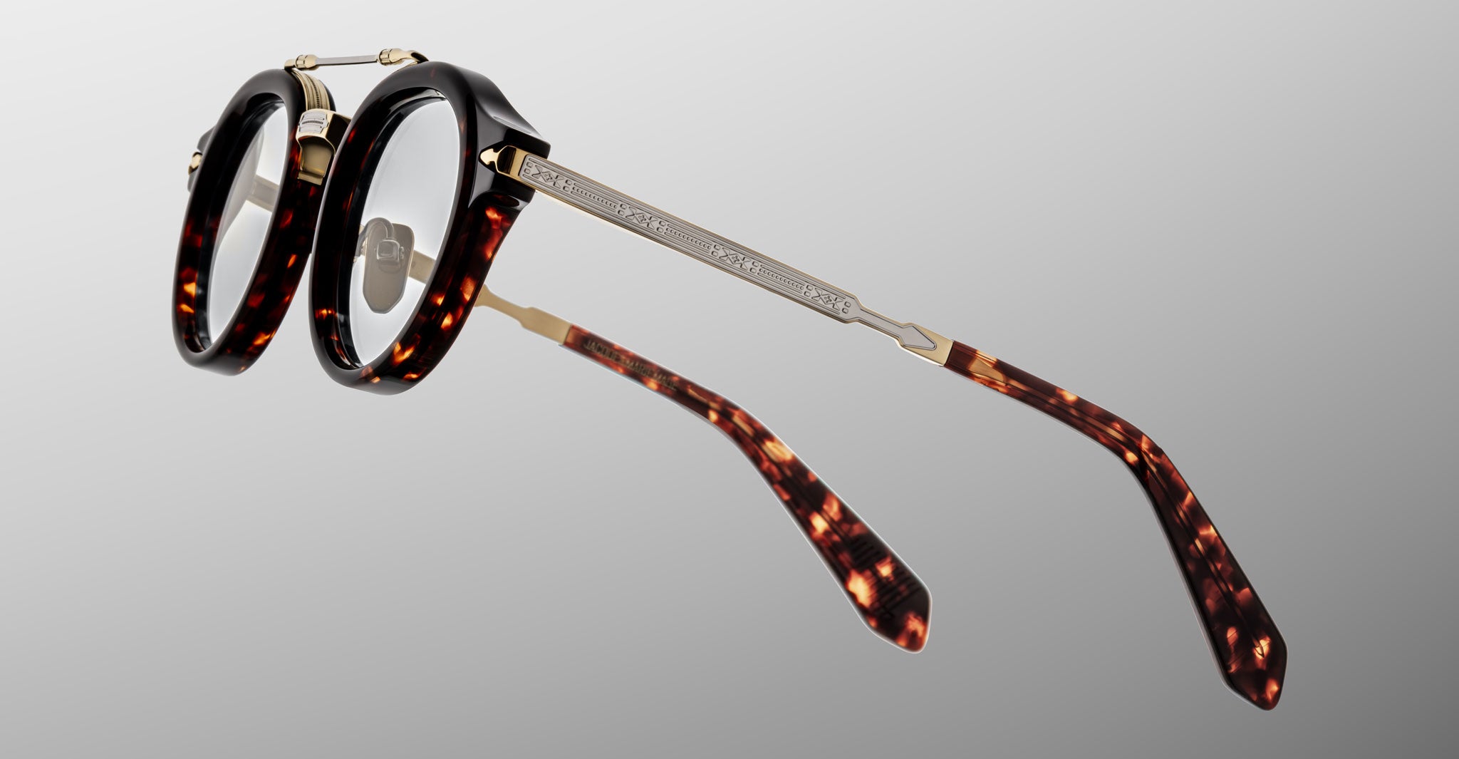 The Jacques Marie Mage Delage features round eyeglasses with thick, dark tortoiseshell frames and gold metal temples, shown against a gray gradient background.