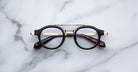 The Jacques Marie Mage Delage eyeglasses, featuring round black rims, gold accents, and tortoiseshell arms, rest on a white marble surface.