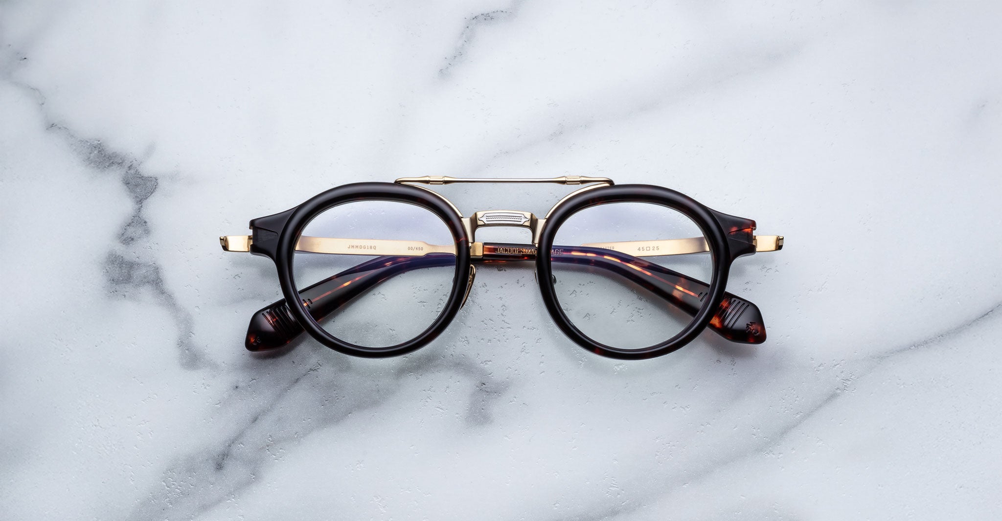 The Jacques Marie Mage Delage eyeglasses, featuring round black rims, gold accents, and tortoiseshell arms, rest on a white marble surface.
