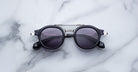 The Jacques Marie Mage Delage, a pair of round black sunglasses with dark lenses and silver accents, rests on a white marble surface featuring subtle grey veining.