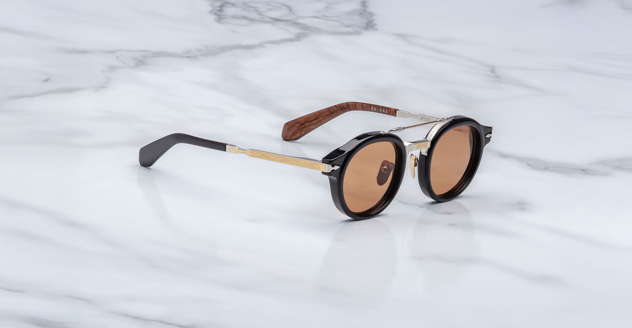 The Jacques Marie Mage Delage are round sunglasses featuring black frames, brown lenses, gold metal arms, and dark brown temples, displayed on a white marble surface.