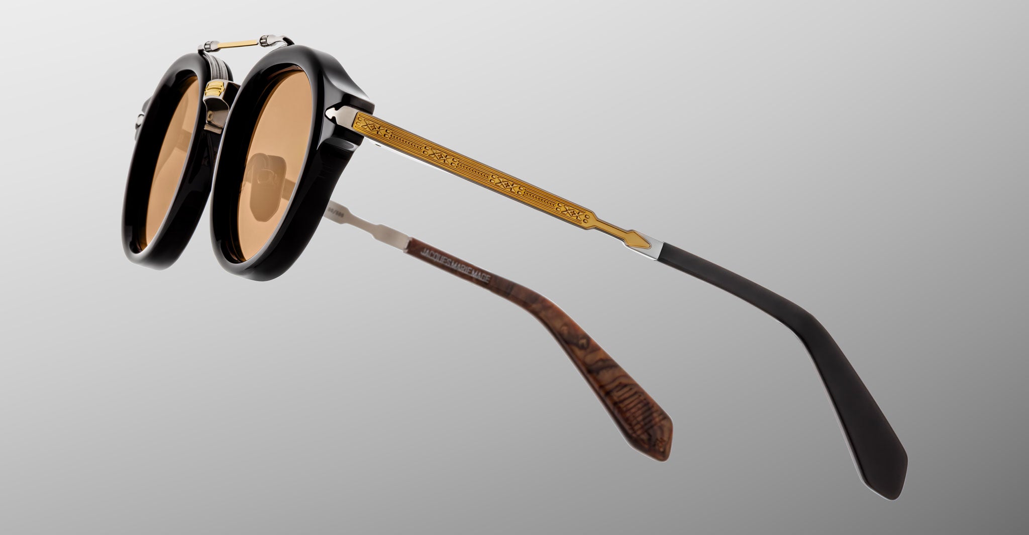 The Jacques Marie Mage Delage sunglasses feature round black frames, brown-tinted lenses, and gold metal arms with engraved designs, displayed against a plain gray background.