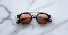 The Jacques Marie Mage Delage sunglasses, featuring a black round frame with brown-tinted lenses and silver accents, rest on a white marble surface.
