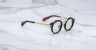 The Jacques Marie Mage Delage eyeglasses, featuring round black rims, gold metal temples, and brown accents, rest on a white marble surface.