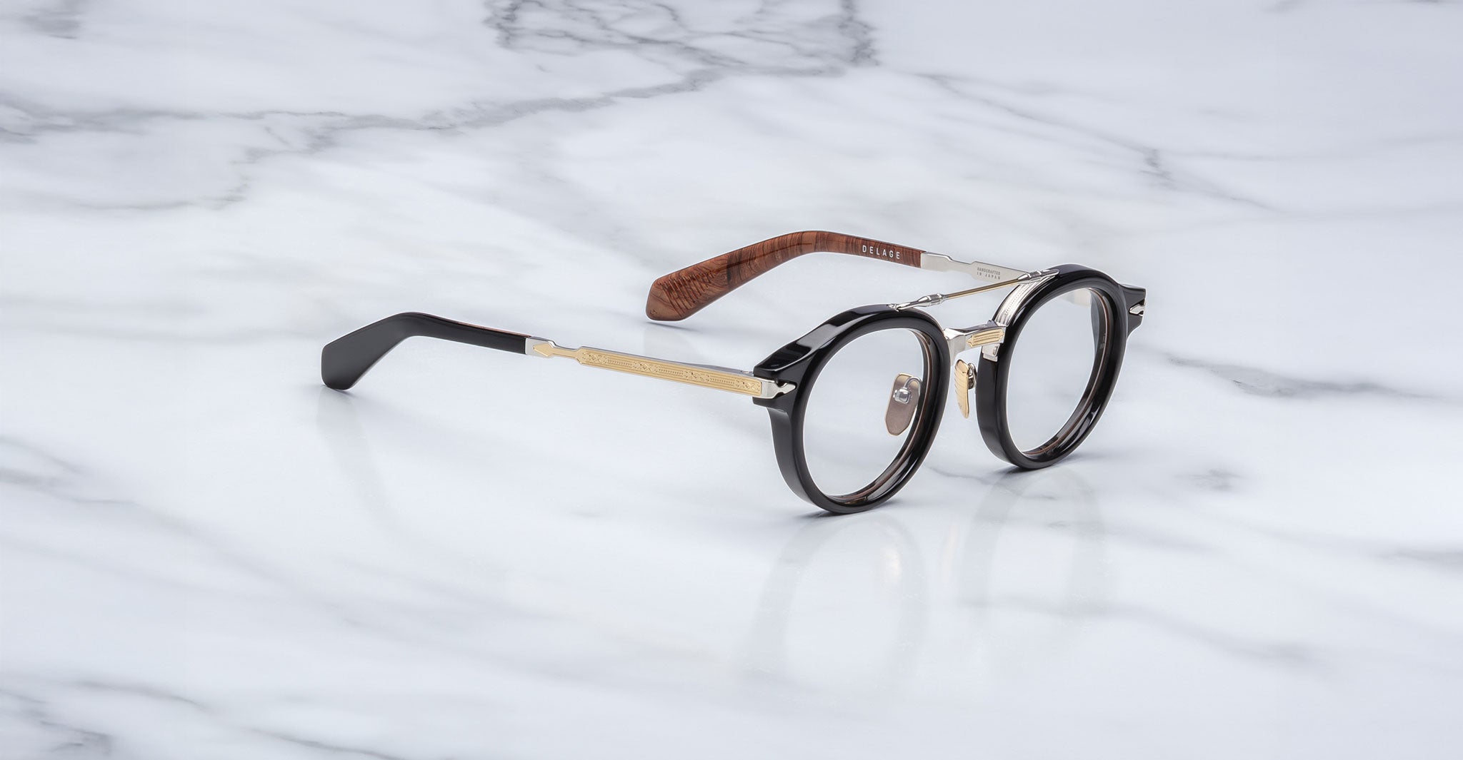 The Jacques Marie Mage Delage eyeglasses, featuring round black rims, gold metal temples, and brown accents, rest on a white marble surface.