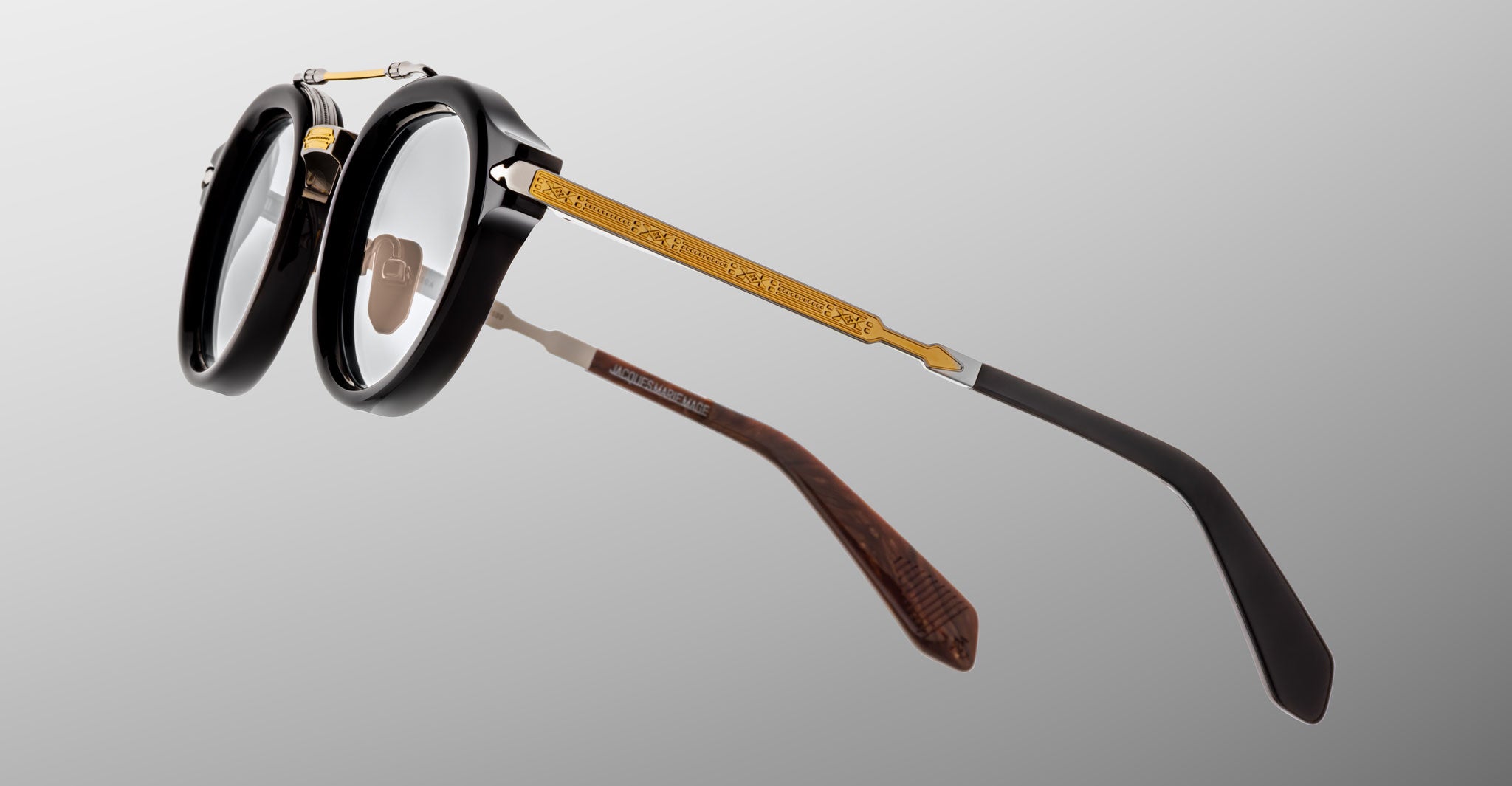 The Jacques Marie Mage Delage features thick black round frames, gold decorative temple detailing, and dark temple tips, displayed on a plain grey background.