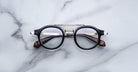 A pair of Jacques Marie Mage Delage round eyeglasses with black rims and brown temples rests on a white marble surface.