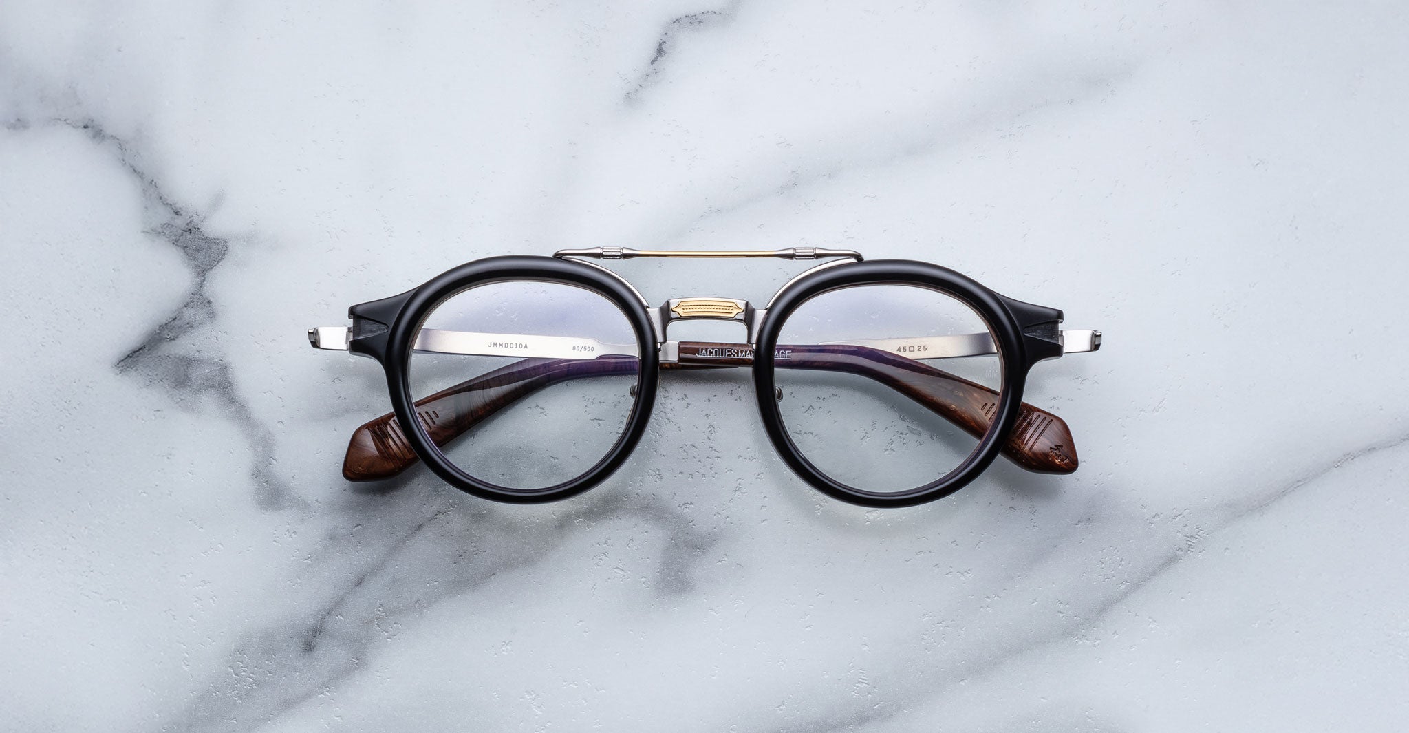 A pair of Jacques Marie Mage Delage round eyeglasses with black rims and brown temples rests on a white marble surface.