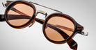 Close-up of Jacques Marie Mage Delage brown tortoiseshell sunglasses featuring round amber-tinted lenses and a metal bridge, set against a light gray background.
