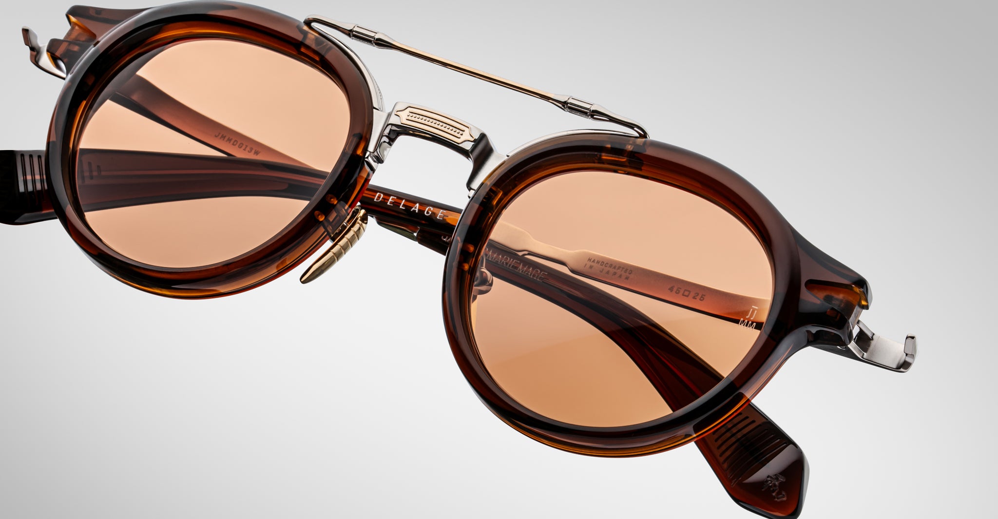 Close-up of Jacques Marie Mage Delage brown tortoiseshell sunglasses featuring round amber-tinted lenses and a metal bridge, set against a light gray background.