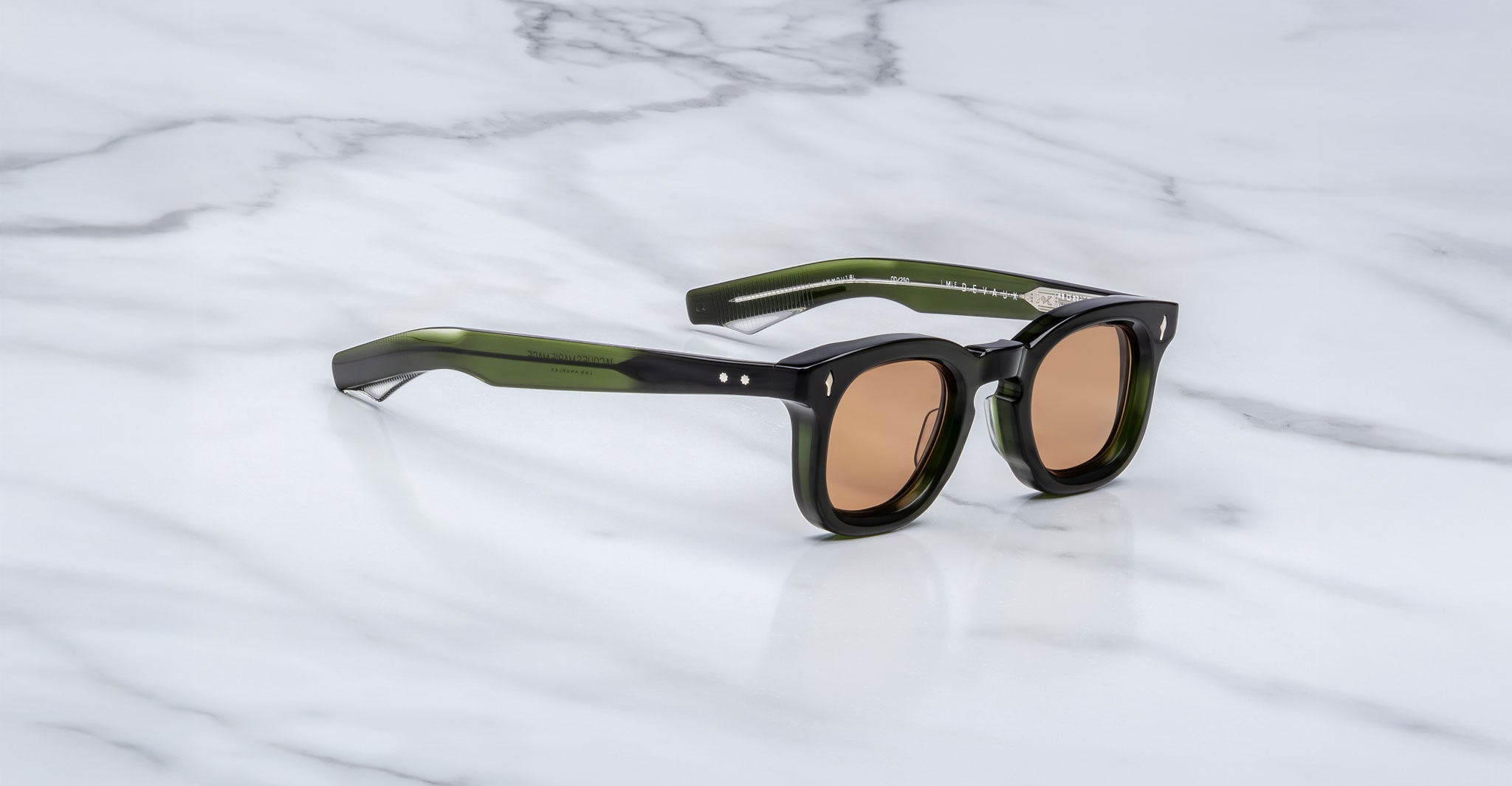 A pair of Jacques Marie Mage Devaux dark green, thick-rimmed sunglasses with brown-tinted lenses rests at an angle on a white marble surface, displaying both the front and side arms.