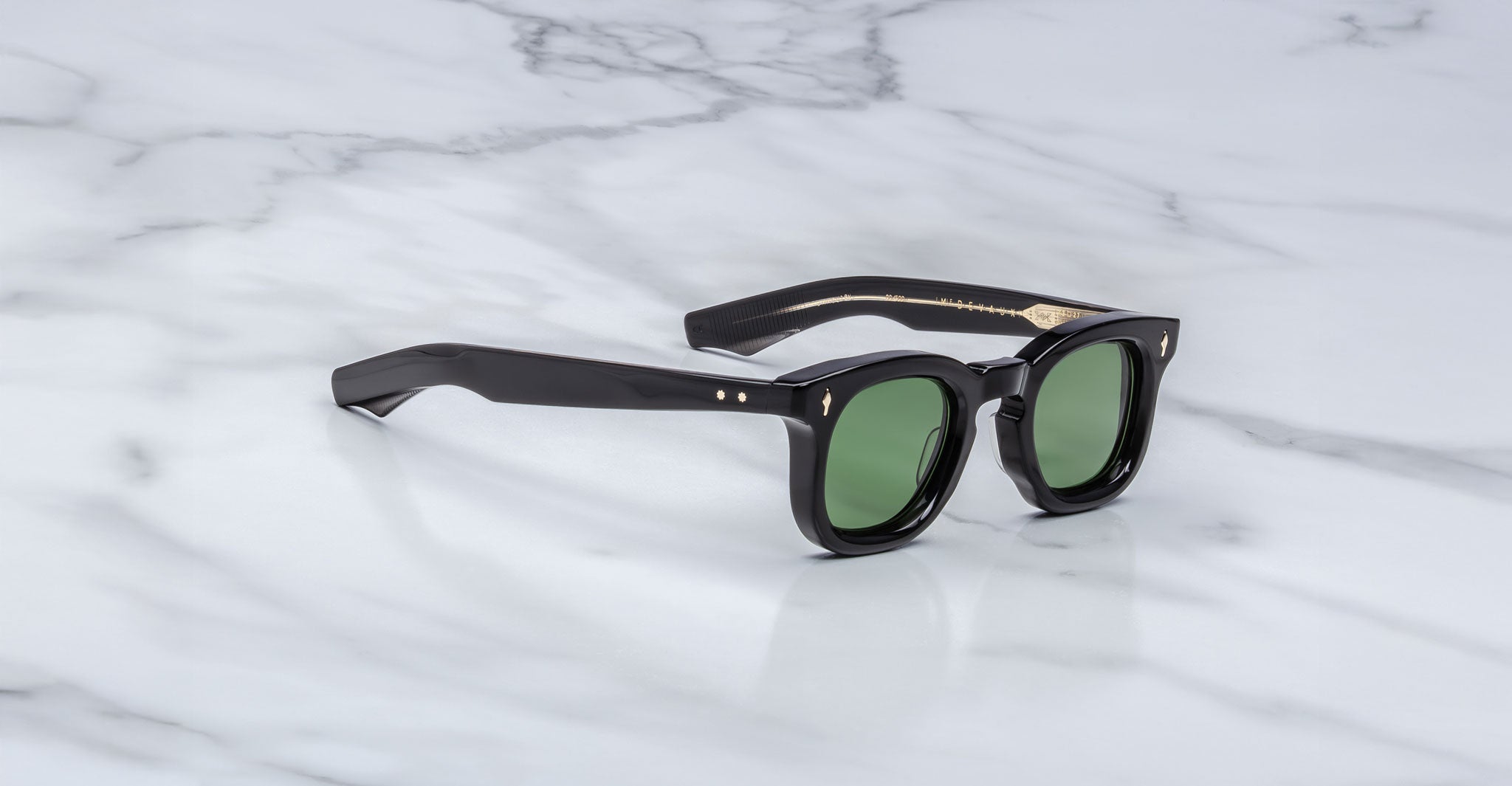 The Jacques Marie Mage Devaux sunglasses, featuring black frames and green-tinted lenses, rest on a white marble surface with grey veining.