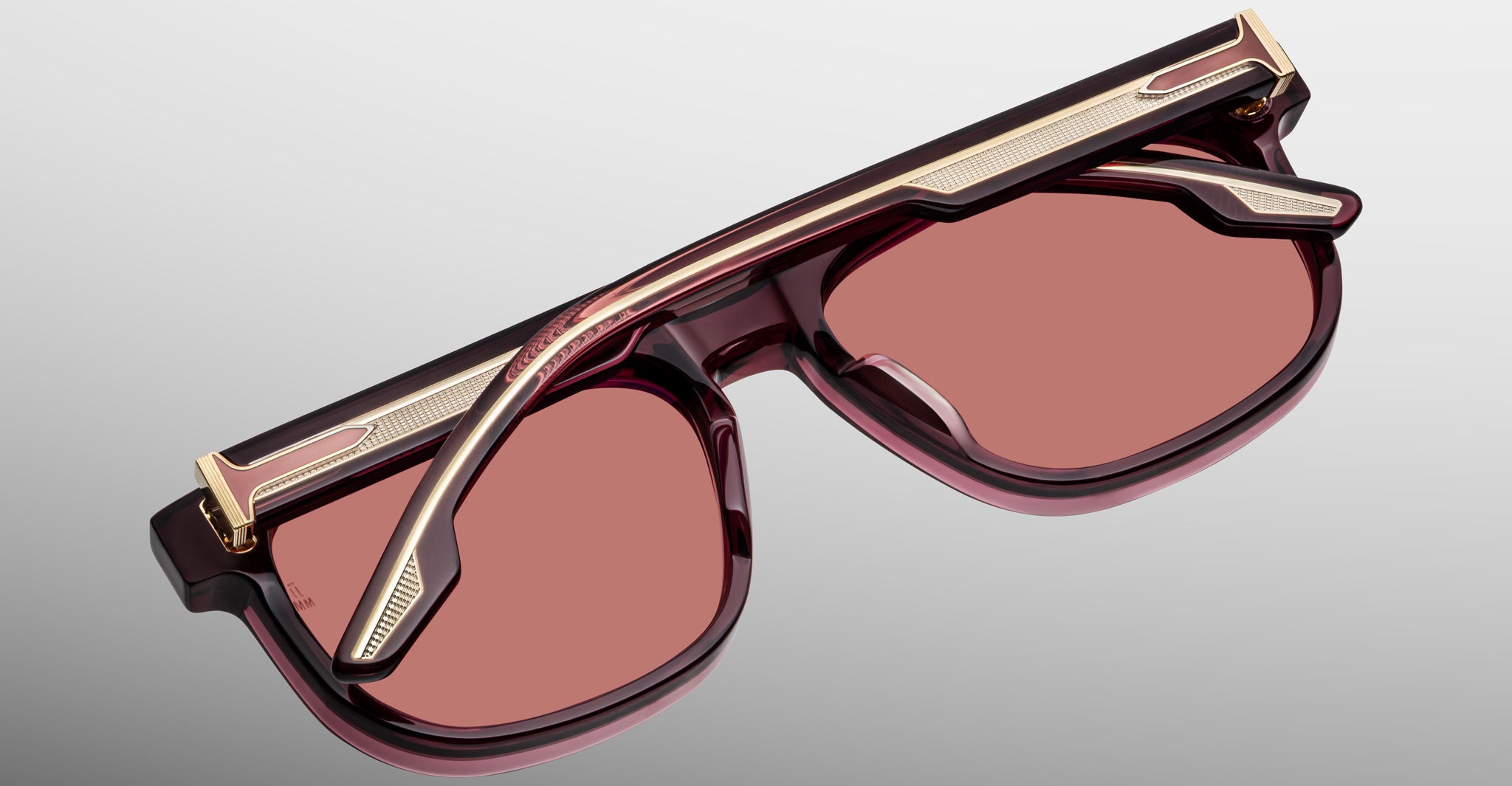 The Jacques Marie Mage Dewitt sunglasses feature rose-tinted lenses, a thick maroon frame, and gold metallic accents, displayed from above with one arm folded across the lenses against a gradient gray background.