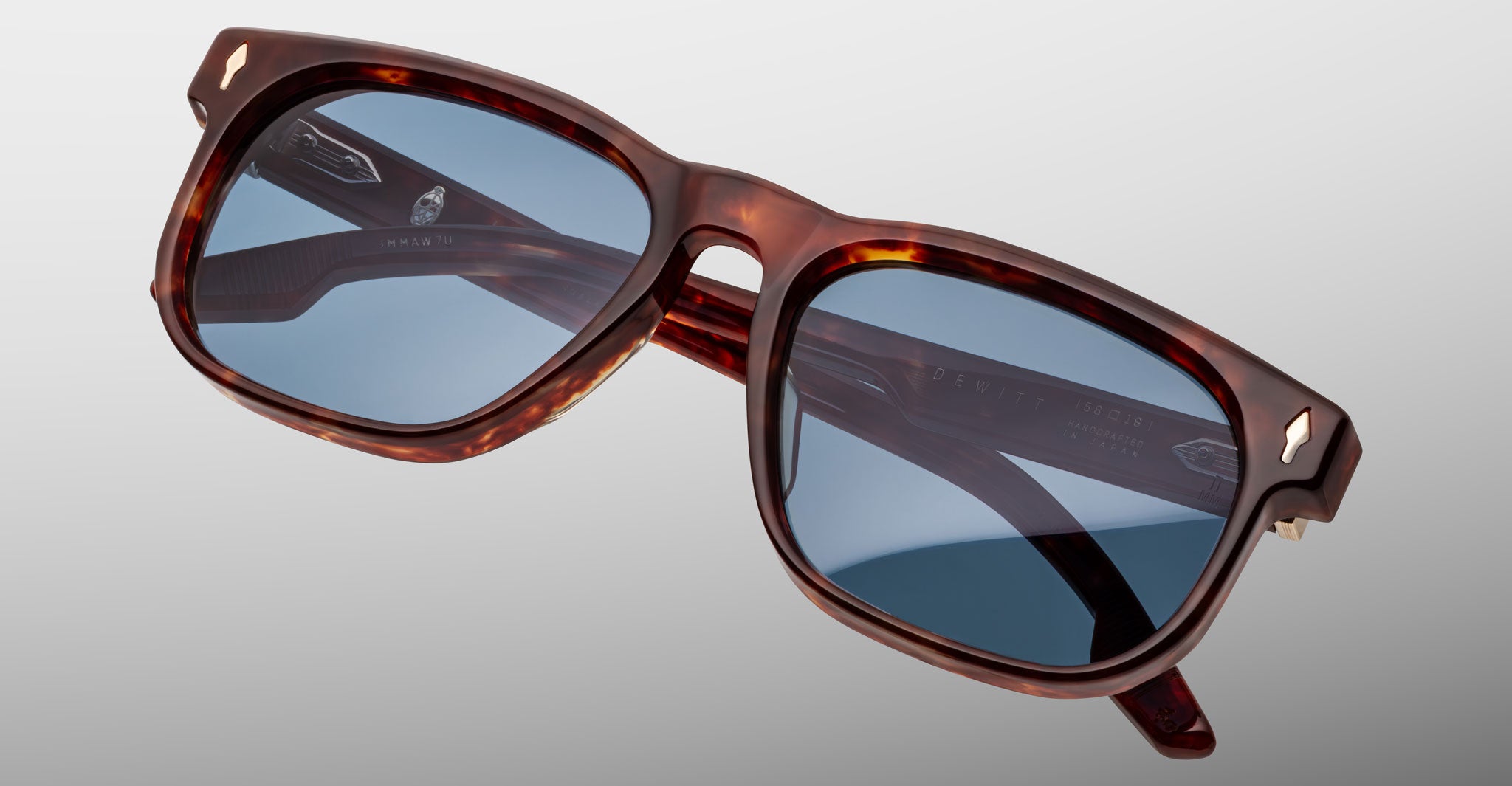 The Jacques Marie Mage Dewitt features rectangular sunglasses with a tortoiseshell brown frame and blue-tinted lenses, set against a plain gray background.