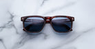 A pair of Jacques Marie Mage Dewitt tortoiseshell sunglasses with dark lenses rests on a white marble surface with gray veining. The arms are slightly open, and the lenses catch some light.