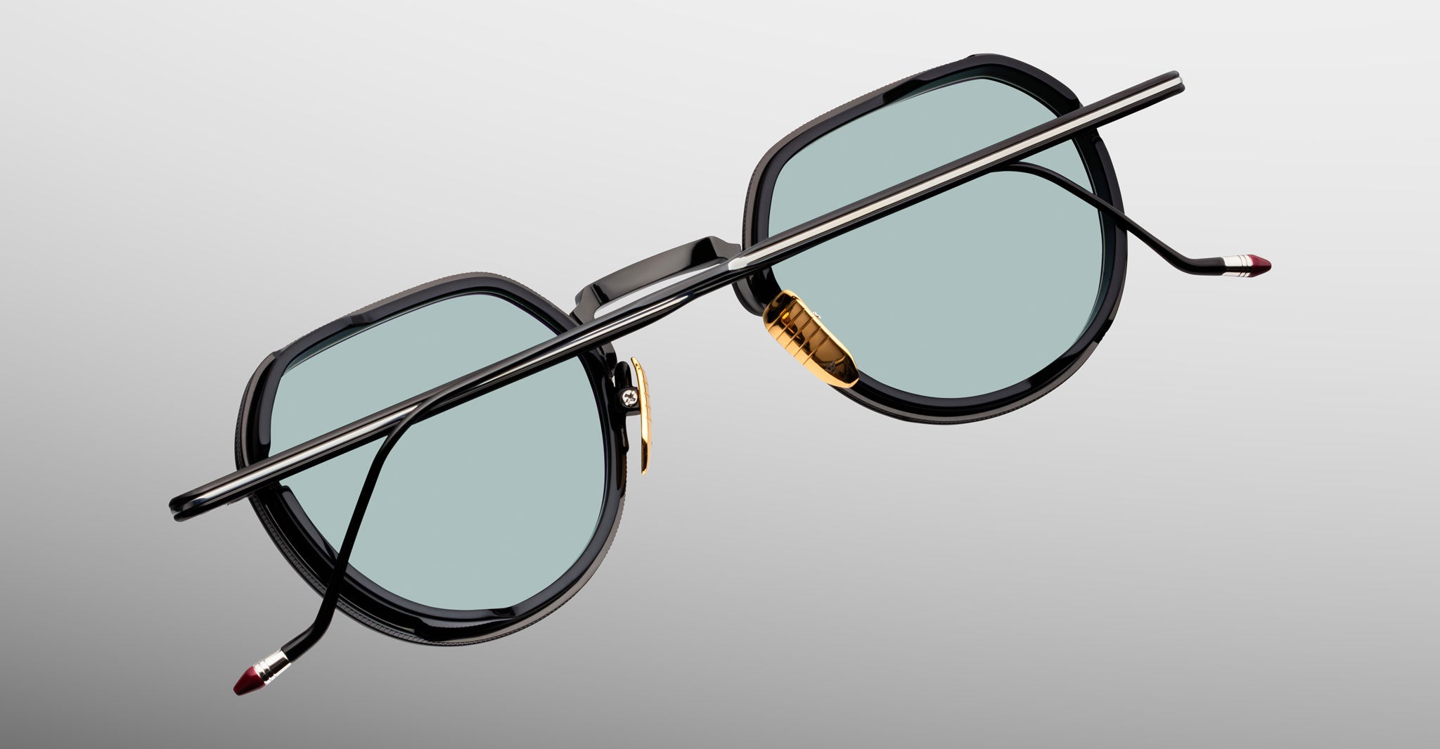 Jacques Marie Mage Dixon sunglasses with black frames, teal lenses, gold nose pads, and thin temples shown from the front.