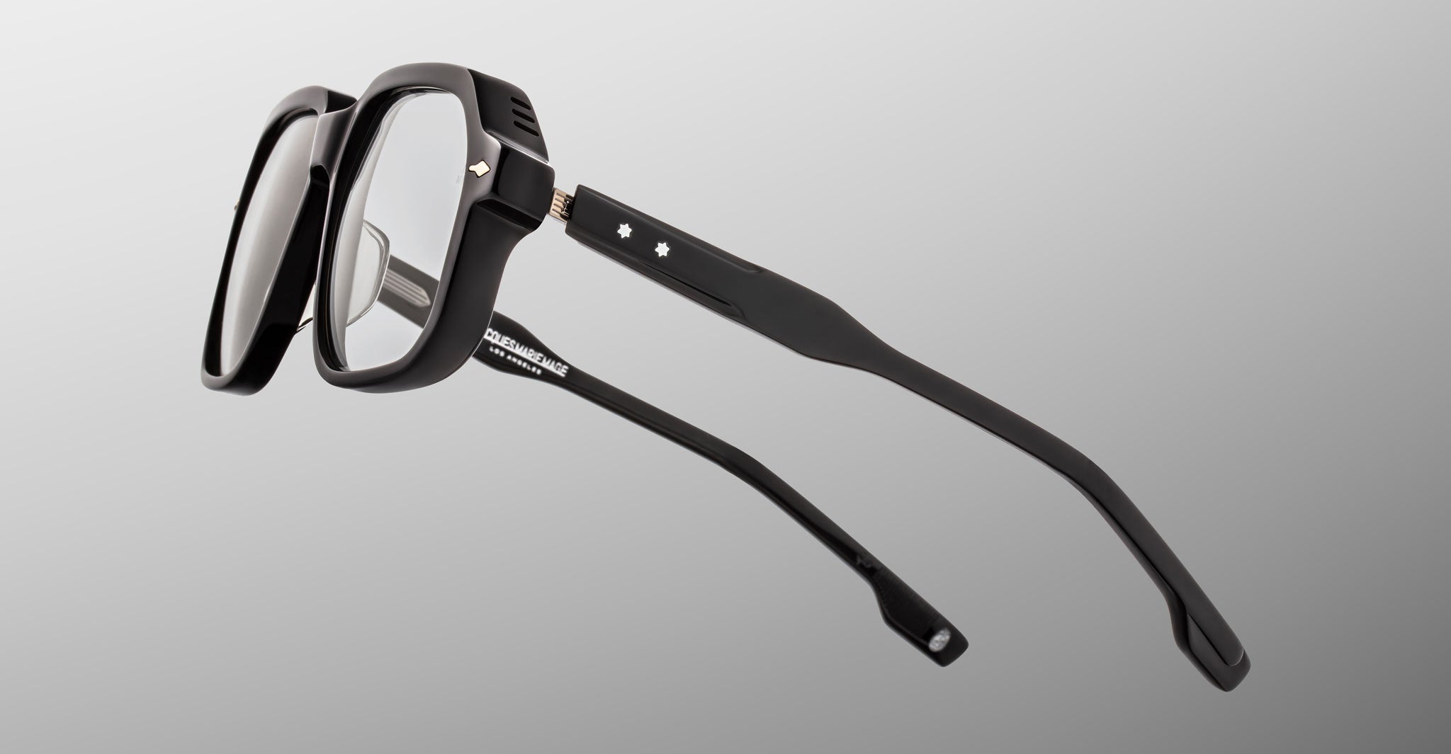 Jacques Marie Mage Domoto: Black rectangular eyeglasses with thick frames and clear lenses, displayed angled on a light gray background. Features white branding inside one arm and small decorative stars on the arms.
