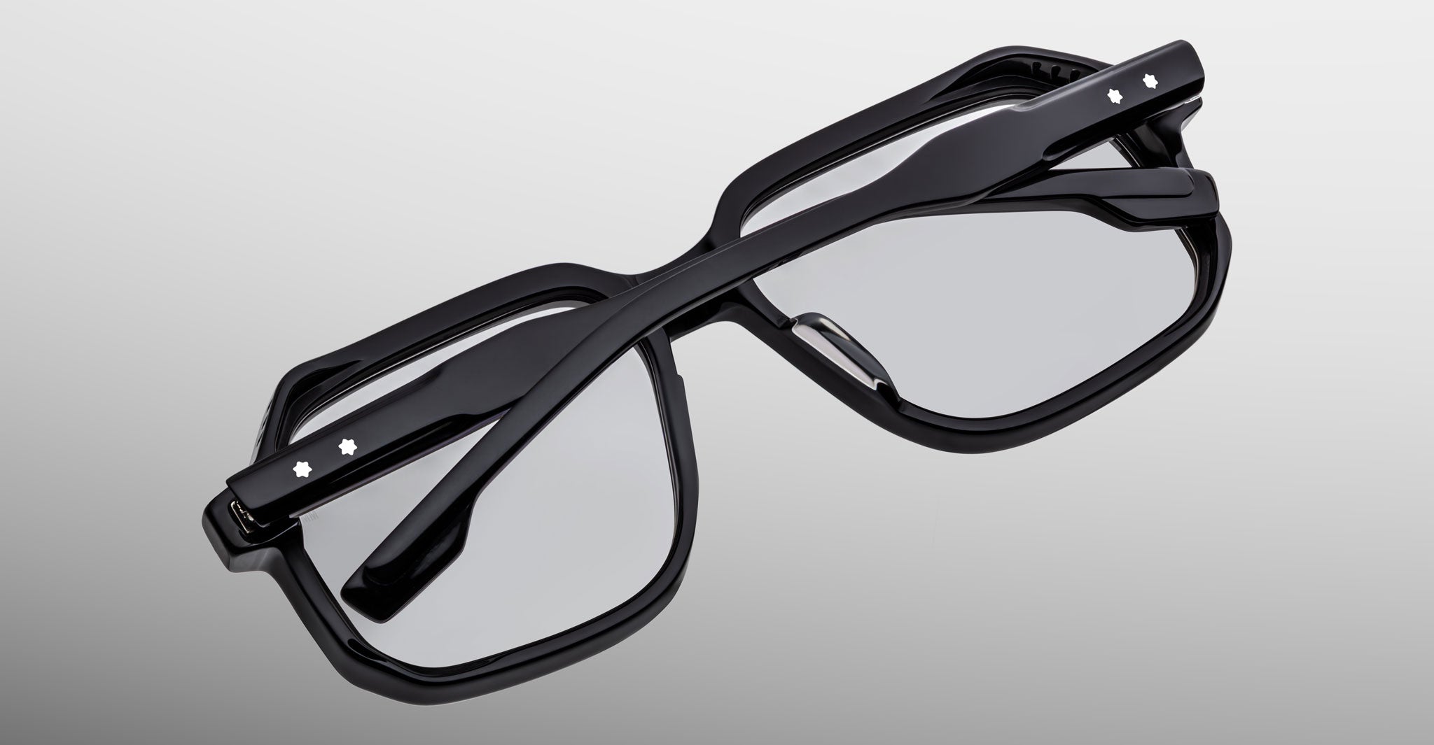 A pair of Jacques Marie Mage Domoto black eyeglasses, both with clear lenses and small white star accents on the temples, handcrafted in Japan, are folded and layered against a light gray background.