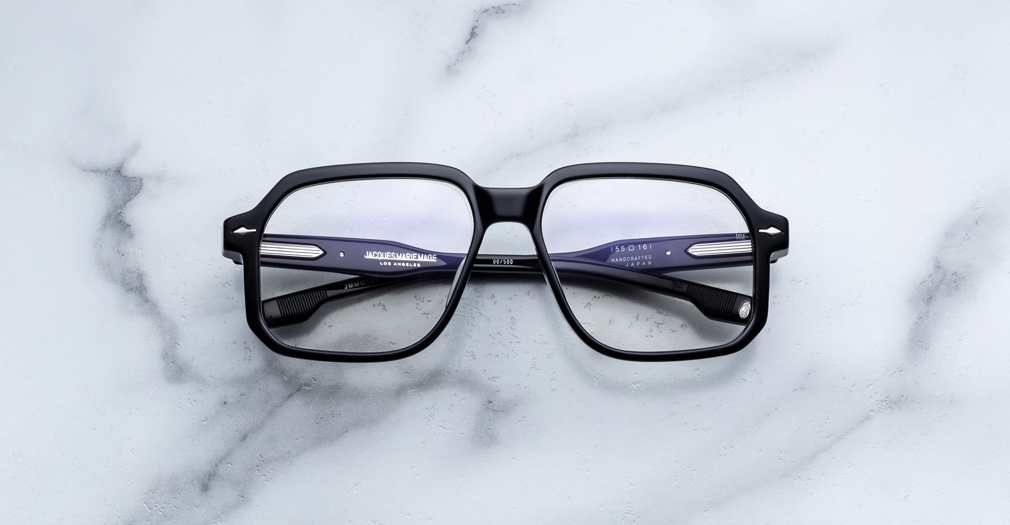 A pair of Jacques Marie Mage Domoto black rectangular eyeglasses, handcrafted in Japan with clear lenses, rests on a white marble surface.