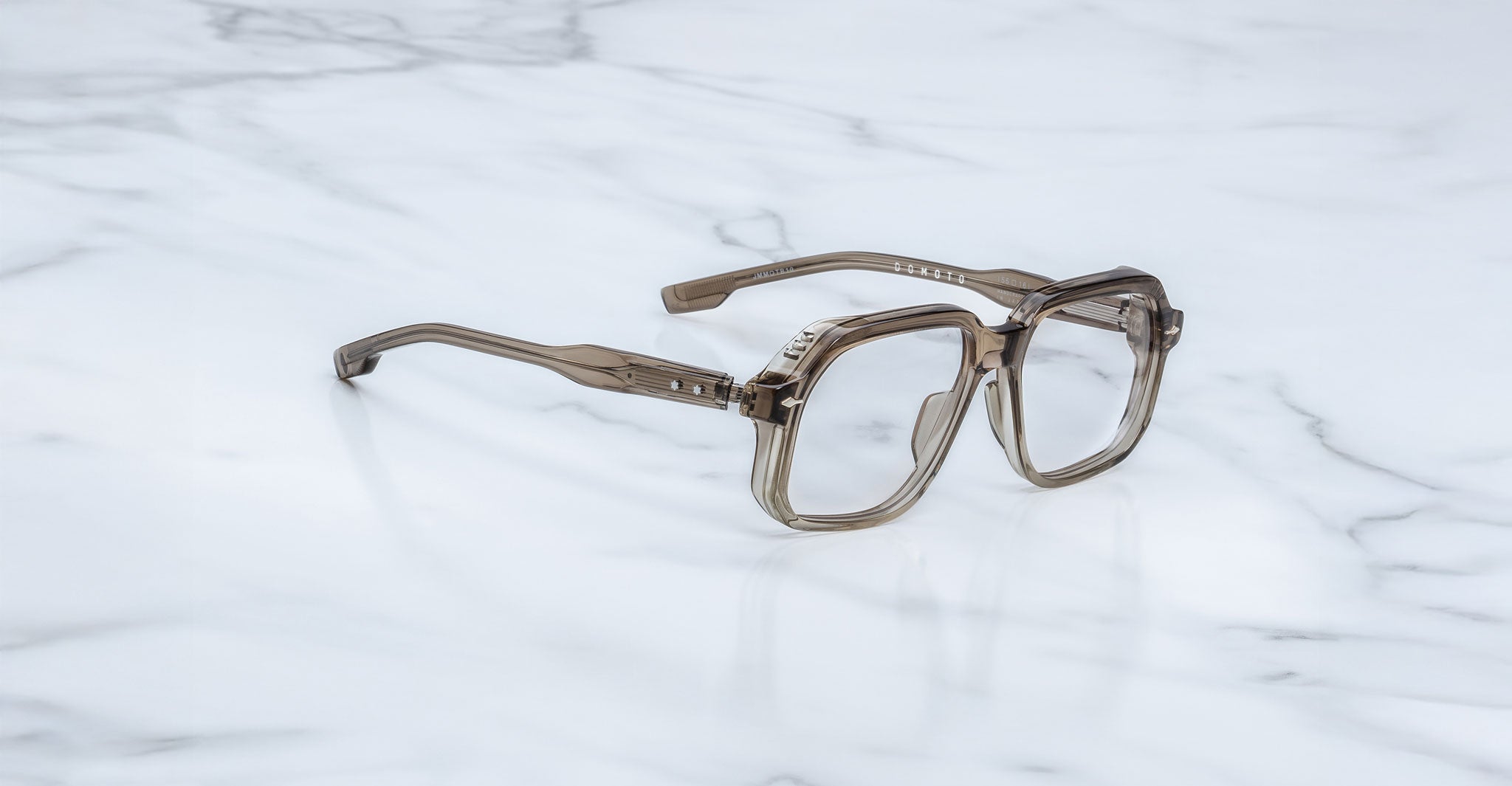 The Jacques Marie Mage Domoto are translucent, square-framed eyeglasses with thick rims and arms, inspired by handcrafted Japanese eyewear, displayed on a white marble surface.