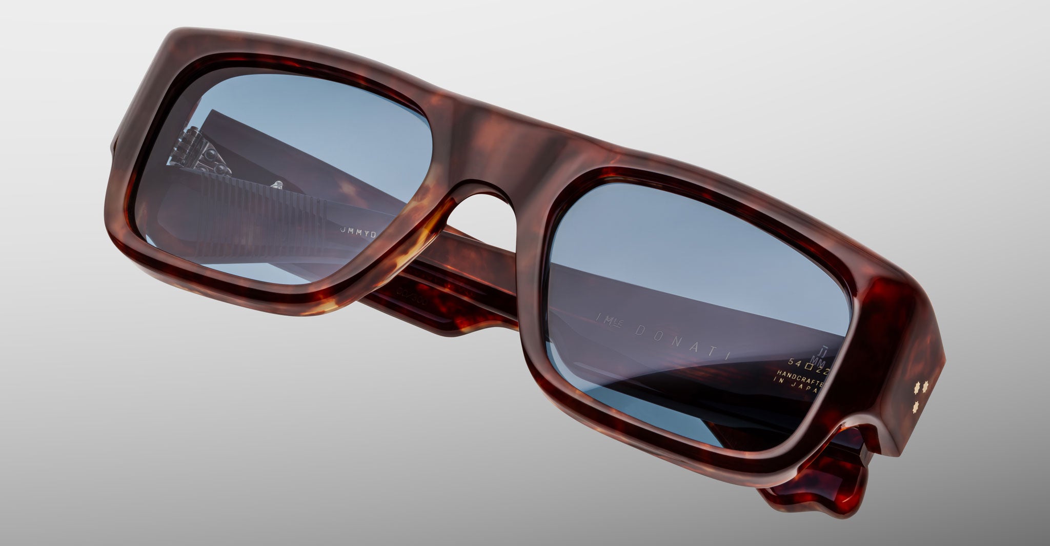 The Jacques Marie Mage Donat features rectangular sunglasses with thick dark tortoiseshell frames and blue-tinted lenses, shown against a plain light gray background.