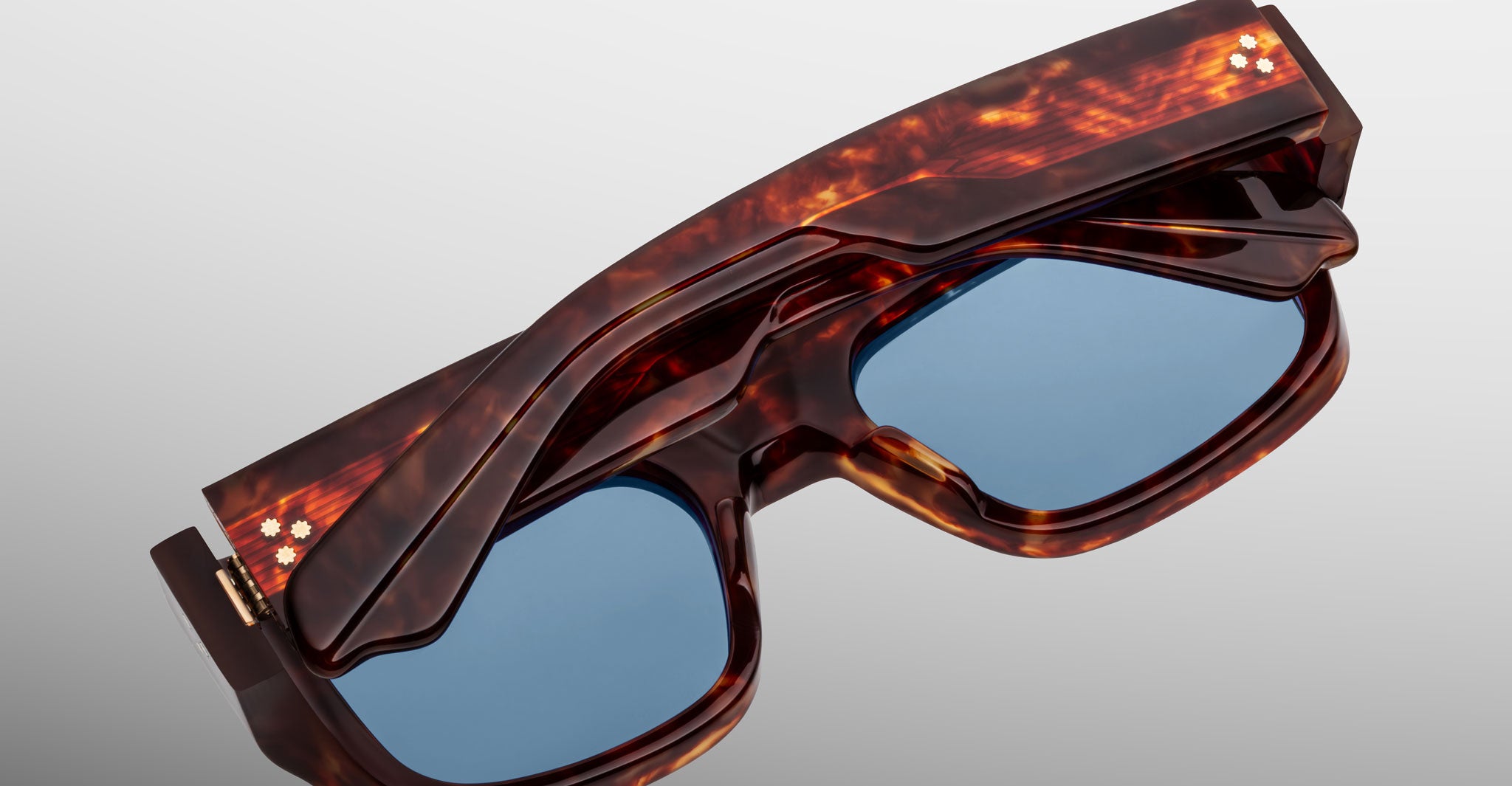 The Jacques Marie Mage Donat sunglasses, featuring a glossy tortoiseshell frame, blue lenses, and two small white dots on the temple hinges, are shown folded neatly against a light grey background.