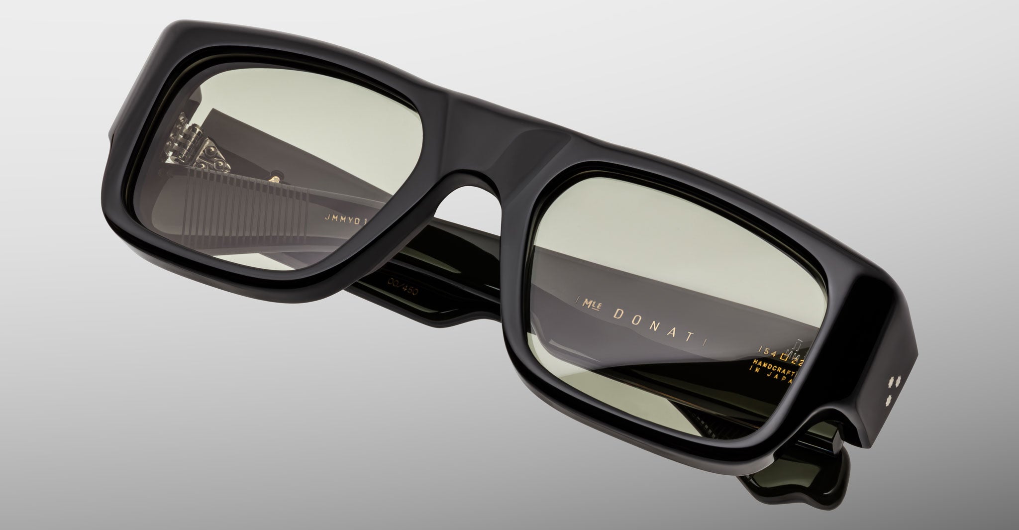 A close-up of Jacques Marie Mage Donat black rectangular eyeglasses with transparent lenses. The arms are folded, showing gold branding text inside the right temple against a light gray background.