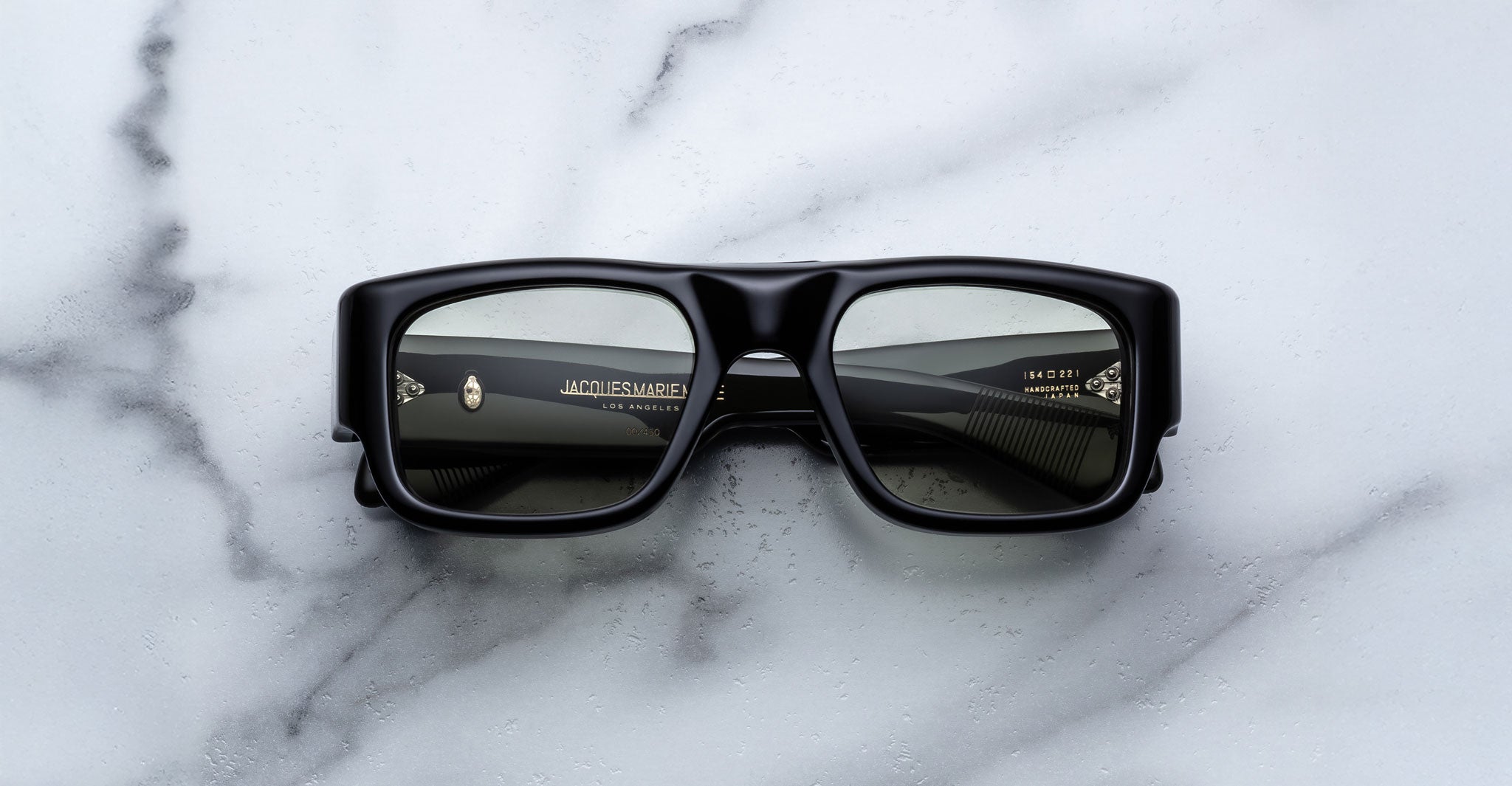The Jacques Marie Mage Donat sunglasses, featuring a rectangular black frame and dark lenses, rest on a white marble surface. White text with brand and sizing details is visible on the inside of the arms.