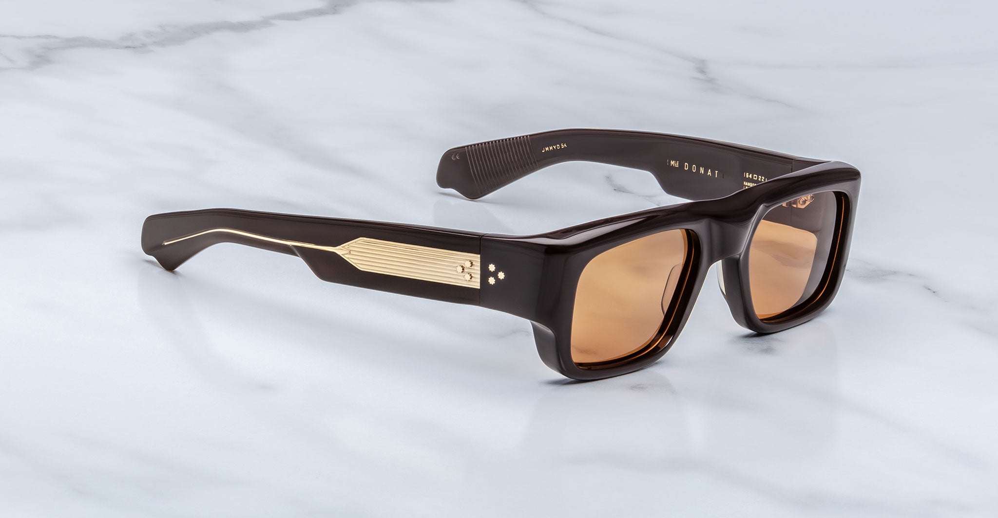 The Jacques Marie Mage Donat sunglasses feature thick, glossy dark brown rectangular frames with gold temple detailing and light brown lenses, displayed on a white marble surface.