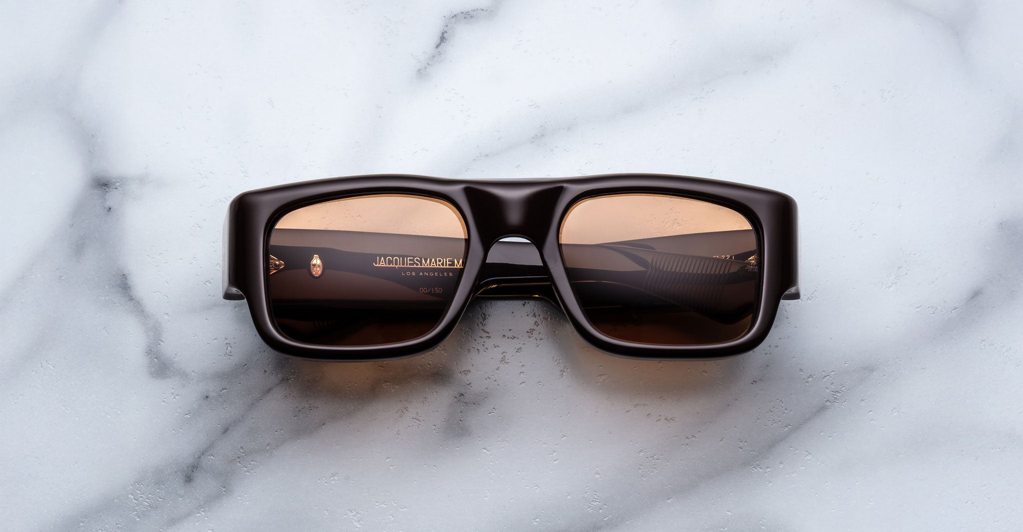 A pair of Jacques Marie Mage Donat dark brown rectangular sunglasses with brown-tinted lenses rests on a white marble surface; the left lens features subtle branding.