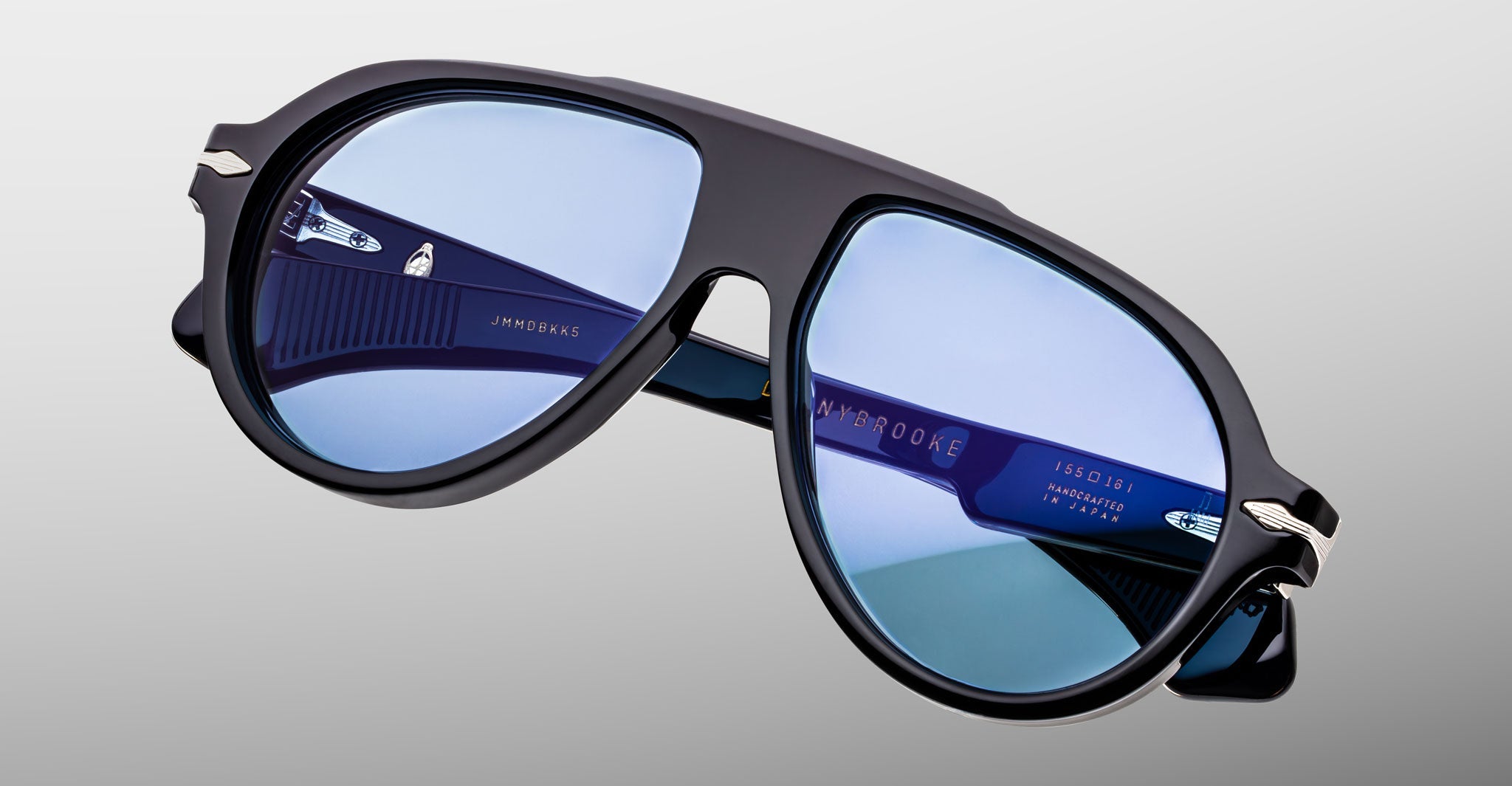 Black aviator-style sunglasses with blue-tinted lenses, viewed at an angle against a plain light gray background. The Jacques Marie Mage Donnybrooke exemplifies the brand’s tradition of refined, handcrafted Japanese eyewear.