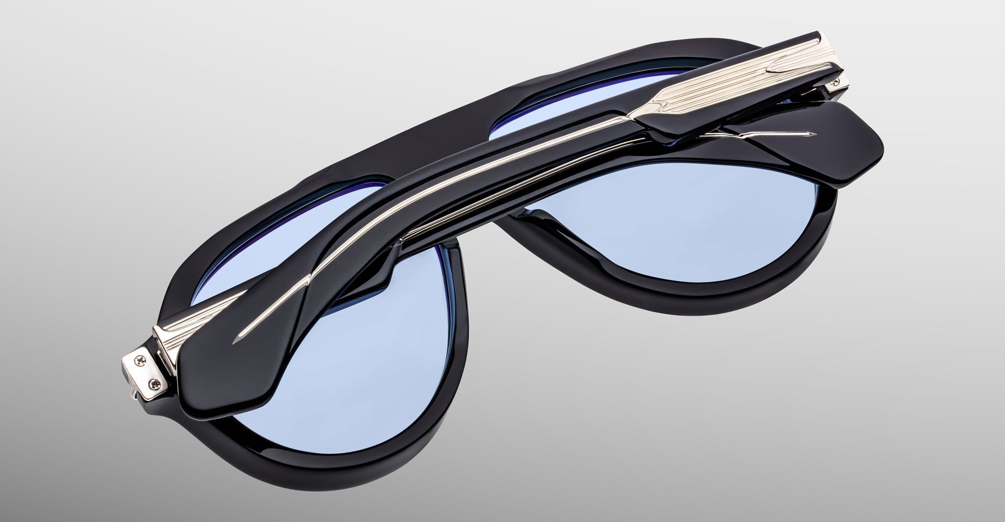 The limited-edition Jacques Marie Mage Donnybrooke sunglasses feature folded black frames, blue-tinted lenses, and silver accents in an oversized oval design, displayed against a light gray background.
