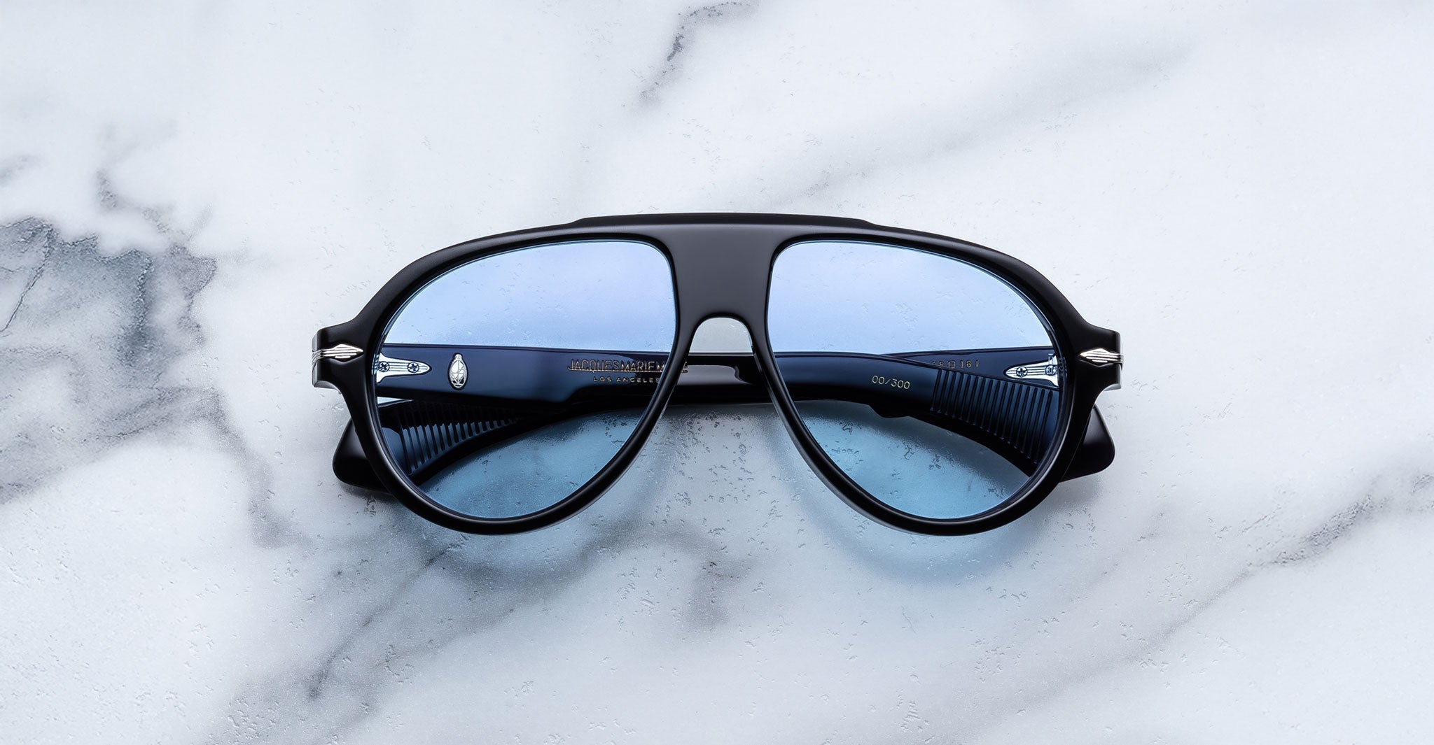 The Jacques Marie Mage Donnybrooke, blue-lensed aviator sunglasses with a black frame inspired by handcrafted Japanese eyewear, are displayed on a white marble surface.