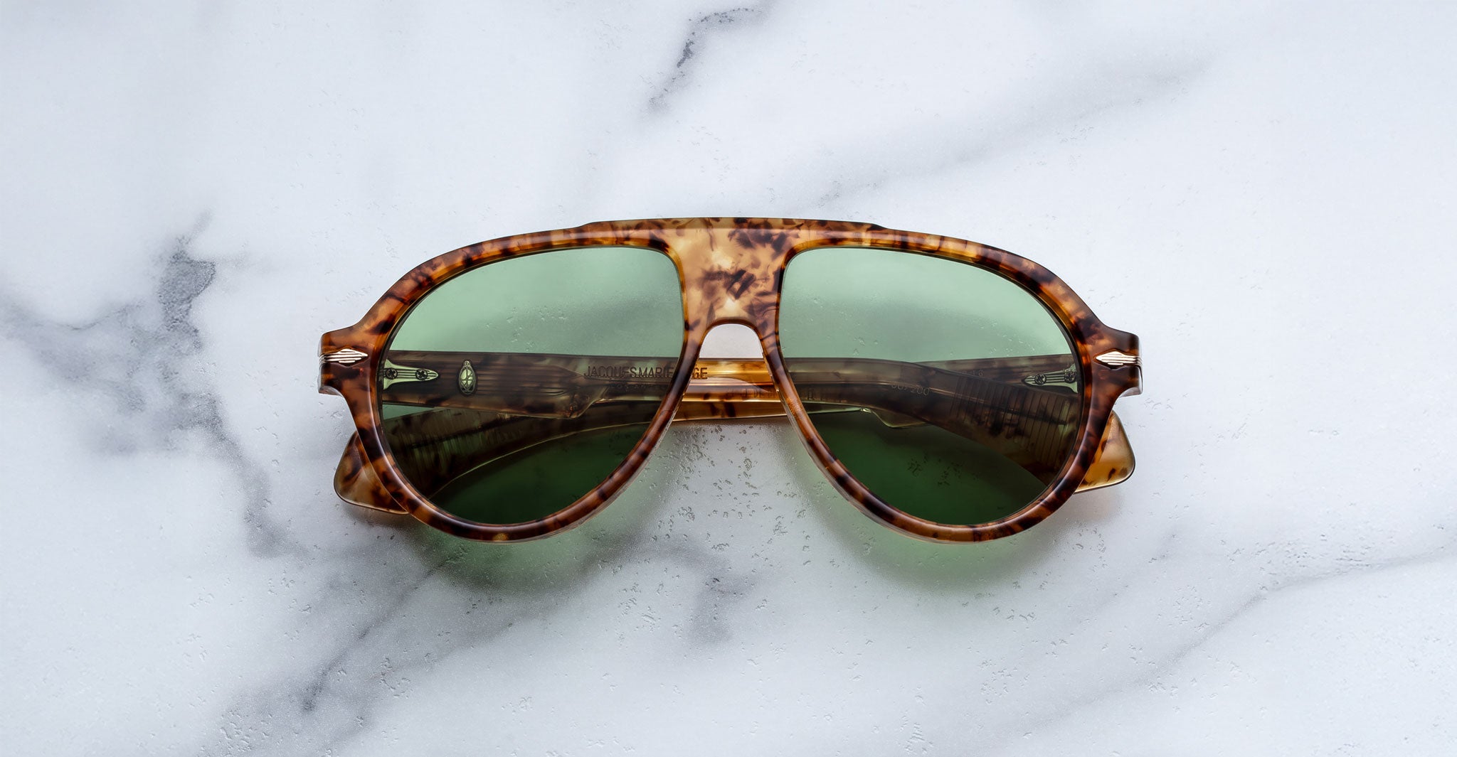 A pair of Jacques Marie Mage Donnybrooke sunglasses featuring tortoiseshell frames and green lenses rests on a white marble surface with gray veining.