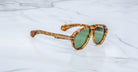 Jacques Marie Mage Donnybrooke sunglasses with green lenses and thick, marbled brown frames rest at an angle on a white marble surface. These limited-edition Jacques Marie Mage frames feature signature tortoiseshell design.