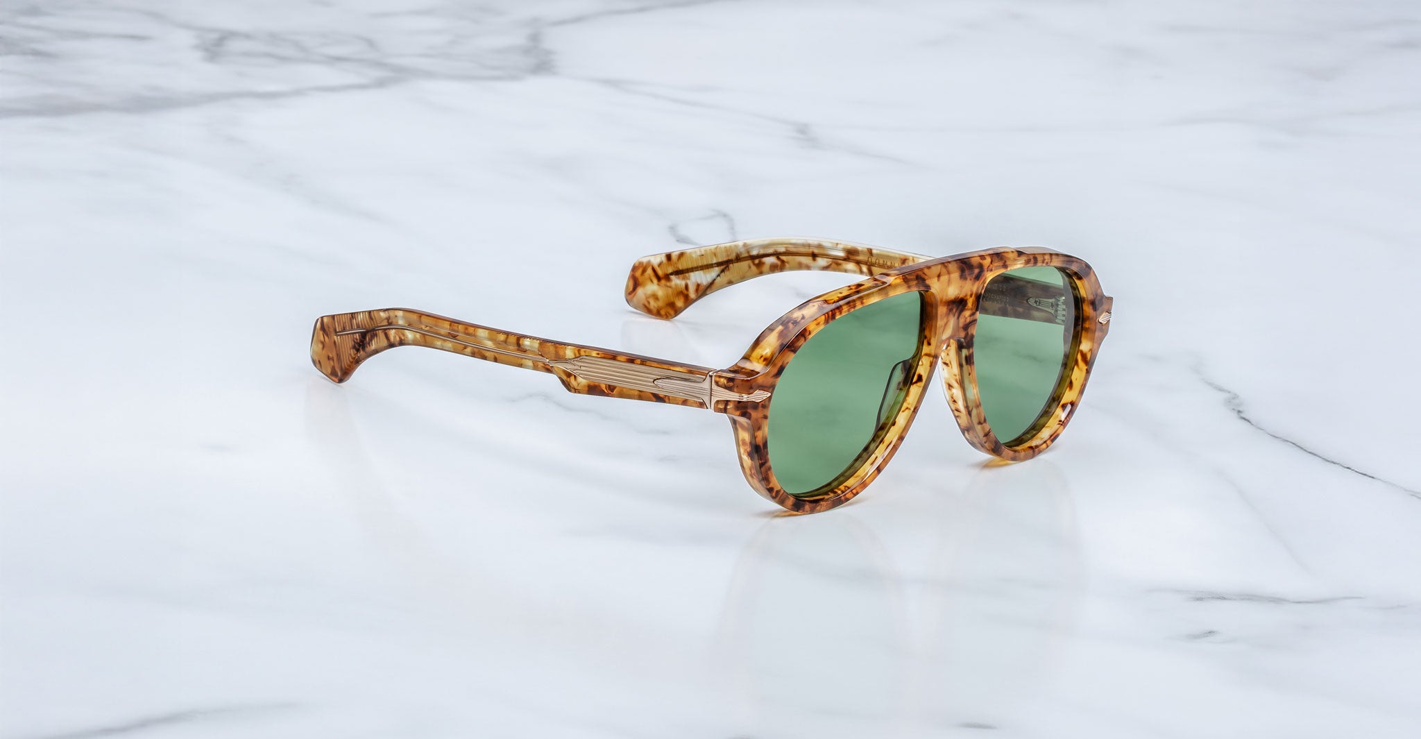 Jacques Marie Mage Donnybrooke sunglasses with green lenses and thick, marbled brown frames rest at an angle on a white marble surface. These limited-edition Jacques Marie Mage frames feature signature tortoiseshell design.