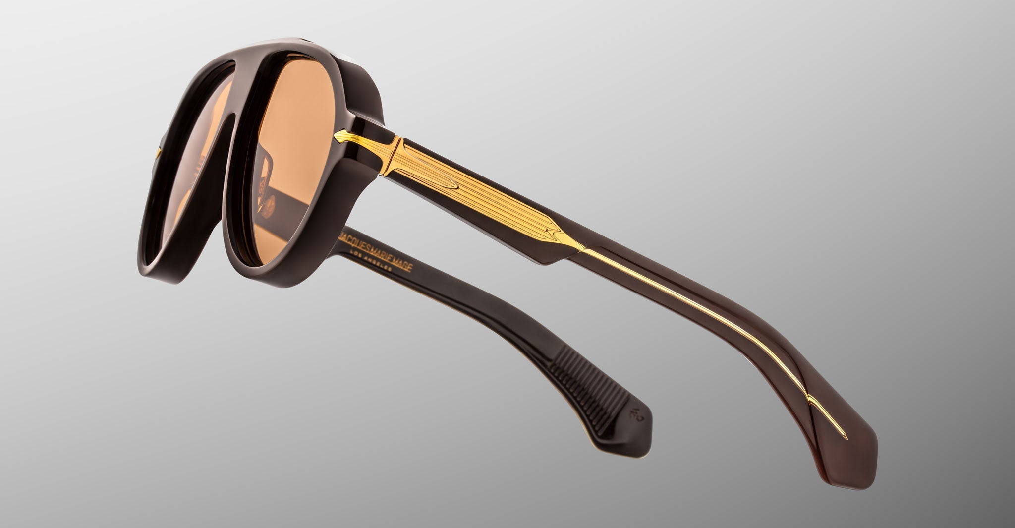The Jacques Marie Mage Donnybrooke sunglasses in brown feature gold metal arm detailing and are shown from a rear side angle against a plain grey background—limited-edition frames that showcase refined craftsmanship.